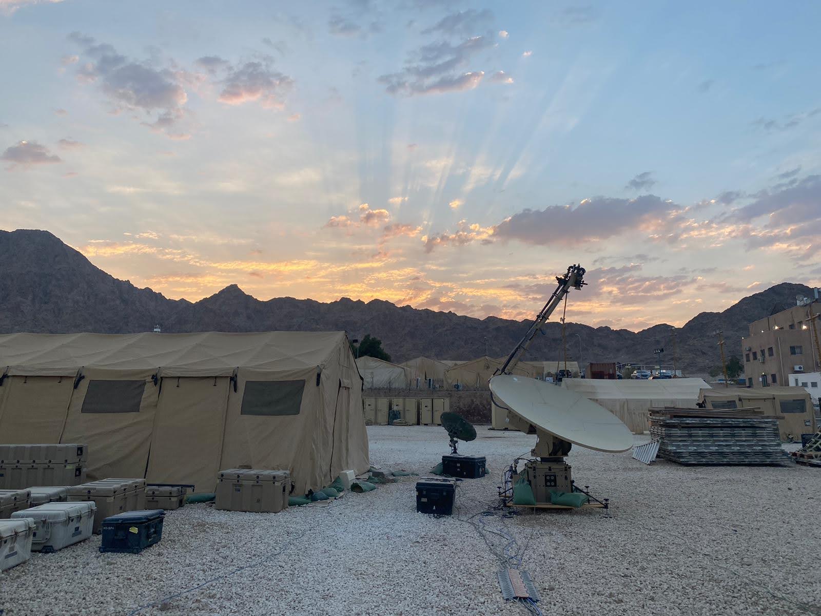 Expeditionary tents and a satellite communications terminal set up in the desert at sunrise, with mountains in the background.